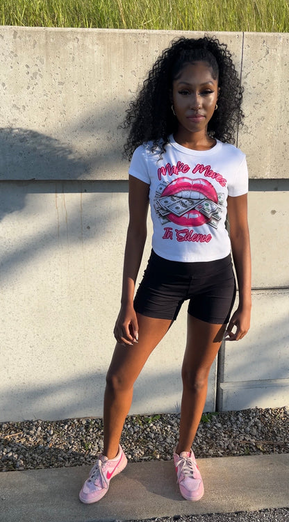 A woman stands outdoors in front of a dark metal wall and wooden steps wearing the Make Moves in Silence women’s baby tee, a white women’s graphic T-shirt featuring a bold lips-and-money graphic with the words “Make Moves in Silence.” She pairs the women’s crop top with black shorts and pink and hot pink sneakers.