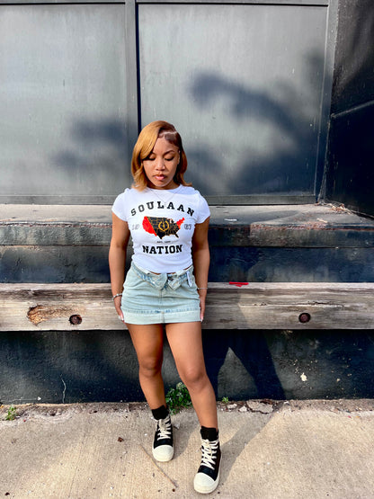 A woman stands outdoors in front of a dark metal wall and wooden steps, wearing a white Soulaan Signature Women’s Baby Tee that reads “SOULAAN NATION” and features a red-and-black United States map with a gold outline, gold sword, and gold wreath, paired with a light denim skirt and black-and-white high-top sneakers.