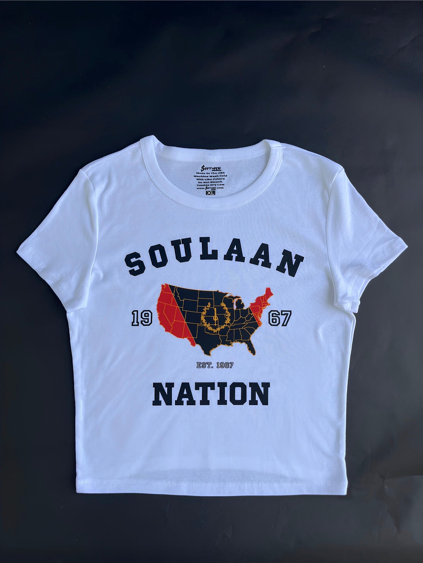 White Soulaan Signature Women’s Baby Tee featuring the words “Soulaan Nation” and “1967,” with a red-and-black United States map, gold outline, gold sword, and gold wreath.