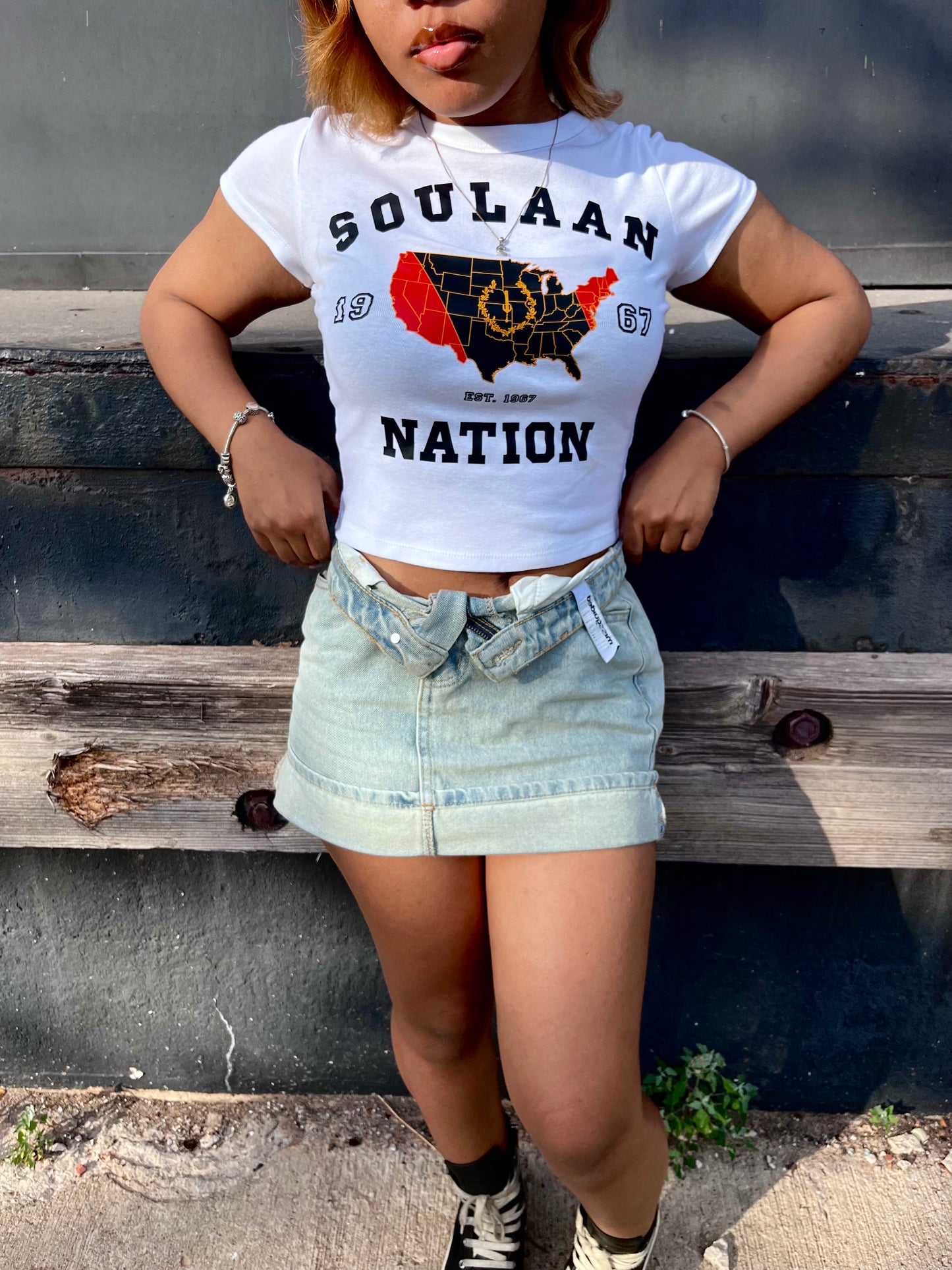 A woman stands outdoors in front of a dark metal wall and wooden steps, wearing a white Soulaan Signature Women’s Baby Tee that reads “SOULAAN NATION” 1967 and features a red-and-black United States map with a gold outline, gold sword, and gold wreath, light denim skirt and black-and-white high-top sneakers.
