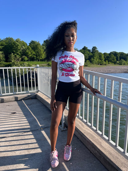 A woman stands on a bridge at the beach with a clear blue sky and trees in the background, wearing the Make Moves in Silence women’s baby tee, a women’s graphic T-shirt featuring bold hot pink lips and a money graphic. She pairs the women’s crop top with black shorts and pink and hot pink sneakers.